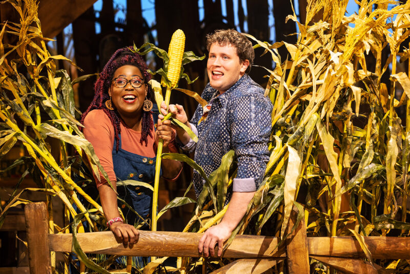 Two people are standing and smiling surrounded by a cornfield with one hand on a wooden fence. They are holding a stalk of corn together in their hands.