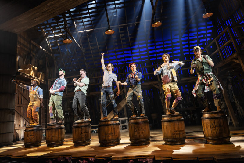 Seven people stand on top of whiskey barrels in the barn. They have lights over them. They are all smiling and the middle character has his hand triumphantly in the air.